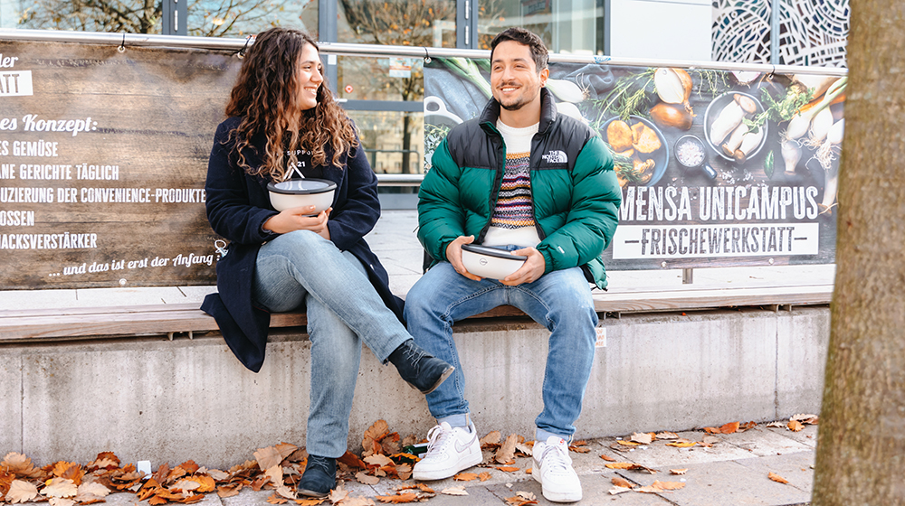 Zwei Studierende sitzen vor der Mensa mit den ToGo-Behältern (Foto: Hannah Theile / Uni Magdeburg)
